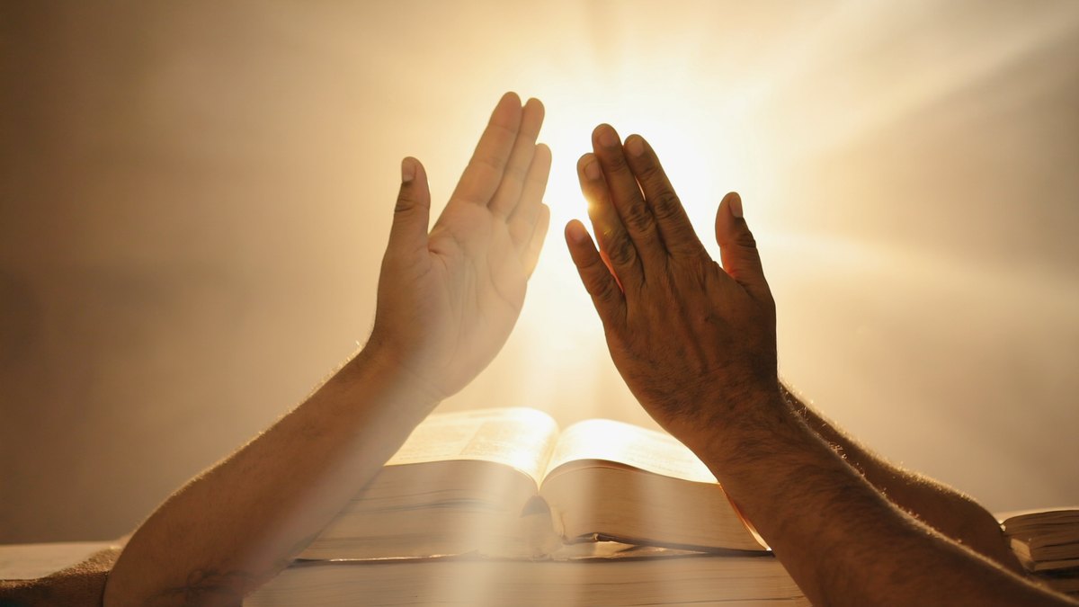 Dos manos abiertas en oración sobre una Biblia abierta, bajo una luz cálida